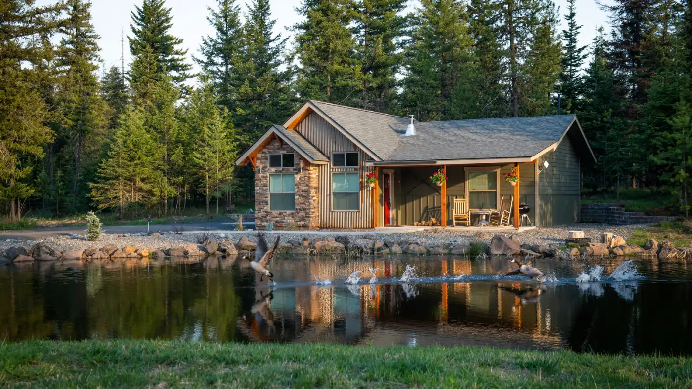 A furnished cabin rental beside a pond, with a covered porch, rocking chairs, a grill, gravel paths and a stone-edged fire pit